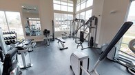 This fitness center offers a comprehensive workout environment designed to accommodate a range of exercise needs. The space is anchored by large, floor-to-ceiling windows that flood the room with natural light and provide open views of the surrounding streetscape, creating an energizing atmosphere for training. The high ceilings enhance the sense of openness and airiness throughout.

The facility is outfitted with a well-rounded selection of equipment, including a multi-station cable machine, adjustable weight benches, a leg press, and a variety of resistance machines suited for both upper and lower body training. Cardio equipment, including a stationary bike, is also present to round out the offering. Full-length mirrors line one wall, supporting proper form and contributing to the overall sense of spaciousness.

The flooring consists of durable, interlocking rubber tiles in a dark tone, providing both safety and comfort underfoot during workouts. The neutral color palette — white walls, gray equipment, and dark flooring — gives the space a clean, modern aesthetic. The single-floor layout ensures all amenities are easily accessible, making this fitness center a practical and well-appointed facility for daily use.