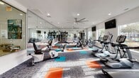 Located in Myrtle Beach, South Carolina, this fitness center is a thoughtfully designed, single-floor facility offering a comprehensive workout environment. The space is defined by its bright, airy atmosphere, thanks to an abundance of natural light streaming through large windows that frame views of the surrounding outdoor amenity areas, including a pool and lush landscaping.

The interior features a contemporary aesthetic with crisp white walls, recessed lighting, and a bold, multi-toned carpet featuring a dynamic pattern of teal, orange, and gray — adding energy and visual interest to the space. Large ceiling fans with brushed metal finishes provide comfortable airflow throughout.

The fitness floor is well-organized and generously proportioned, accommodating a wide variety of equipment. Cardio machines include multiple treadmills, stationary bikes, ellipticals, and a rowing machine. A dedicated free-weight area features a full rack of dumbbells, an adjustable bench, and a cable/multi-use strength machine. Floor-to-ceiling mirrors line key sections of the walls, enhancing the sense of space and supporting form-focused training.

Multiple flat-screen televisions are mounted throughout, and colorful wall art with a coastal bicycle theme ties the space to its beachside locale. The overall ambiance is modern, motivating, and well-suited for a full range of fitness activities.
