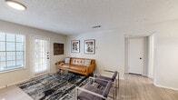 This thoughtfully designed one-bedroom, one-bathroom residence offers a comfortable and functional layout across a single floor, encompassing a living room, bedroom, bathroom, closet, laundry area, and hallway. The living room serves as the heart of the home, showcasing a striking white brick fireplace as its focal point, complemented by hardwood-style flooring, a front entry door with a sidelite window, and natural light filtering through multiple windows. The open layout connects seamlessly to the rest of the unit. The bedroom features plush carpeting, a ceiling fan with light, and a distinctive wood-framed accent wall. Two doorways lead directly to a dedicated walk-in closet with built-in shelving and to the hallway, offering excellent flow and storage. The full bathroom is well-appointed with a dual-sink vanity featuring a light stone countertop, matte black fixtures, a large mirror, and a tub-shower combination. A dedicated in-unit laundry area with an included top-load washer and overhead shelving adds everyday convenience. The textured ceilings and neutral color palette throughout lend the space a clean, cohesive aesthetic.
