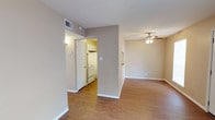 This single-floor residence offers a clean, open layout designed for comfortable everyday living. The main living area is anchored by warm hardwood-style flooring that extends throughout, creating a cohesive and inviting atmosphere. A ceiling fan with a light fixture serves as a practical centerpiece overhead, providing both airflow and ambient lighting to the space.

Neutral, warm-toned walls in a soft taupe palette complement the flooring and contribute to a bright, welcoming feel. The open flow between the living area and the adjacent spaces allows for flexible use and easy movement throughout the unit.

The kitchen is partially separated by a structural wall, creating a semi-defined cooking space while maintaining a connection to the rest of the home. It features white cabinetry and white appliances, including a range, offering a clean and functional setup. A door within the kitchen area provides additional access, adding convenience to the layout.

Ceiling vents are visible throughout, indicating a central HVAC system for year-round climate control. White baseboards frame the flooring neatly, adding a finished, polished look to each room. Overall, the space presents a straightforward, functional design with consistent finishes that create a harmonious and practical living environment.