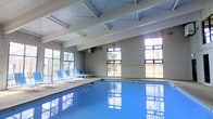 This single-story indoor aquatic facility offers a welcoming and well-lit environment centered around a generously sized swimming pool. The pool's clear blue water is a standout feature, reflecting the abundant natural light that streams in through the large multi-pane windows lining the walls on multiple sides. These expansive windows also provide views of the surrounding tree-lined exterior, connecting the indoor space to the natural environment outside.

The architectural design is defined by a vaulted ceiling with exposed white structural beams, which lend the space an open, airy quality and contribute to an impressive sense of vertical volume. The clean, light-toned walls and painted ceiling reinforce the bright and fresh atmosphere throughout.

A poolside deck area runs along one side of the pool, furnished with blue lounge chairs ideal for relaxation before or after a swim. The overall layout is practical and spacious, providing ample room for both leisure and lap swimming. The facility's design prioritizes natural illumination and openness, making it a comfortable and inviting space year-round. Whether used for recreational swimming or fitness purposes, this indoor pool area combines functional design with a pleasant, light-filled ambiance.