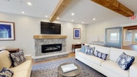 This single-floor common room is thoughtfully designed to encourage gathering and relaxation, combining functional amenities with warm, inviting aesthetics. Exposed wood beams run throughout the ceiling, serving as a defining architectural element that ties the entire space together. Hardwood flooring extends across all areas, contributing to a cohesive and polished look.

The lounge area serves as the social centerpiece, anchored by a striking floor-to-ceiling stone fireplace with a wood mantel and a wall-mounted flat-screen television above. Comfortable seating, including white and cream sofas accented with navy and patterned throw pillows, creates a relaxed coastal-inspired atmosphere. Nautical artwork and decorative accents reinforce this design theme throughout.

The fully equipped kitchen features light gray shaker-style cabinetry, bright white quartz countertops, stainless steel appliances—including a refrigerator, gas cooktop, wall oven, and range hood—and a spacious island with bar seating, making it well-suited for communal use.

Adjacent to the kitchen, a casual dining area accommodates multiple guests at wood-topped tables with white chairs. Large windows allow generous natural light to fill the space while offering views of the surrounding outdoor environment, enhancing the room's open and airy ambiance.