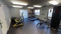 This fitness center is a well-appointed, single-floor exercise facility designed to support a range of workout routines. The room features a dark rubber flooring surface, which provides both durability and safety underfoot during workouts. Overhead, a series of bright fluorescent panel lights ensure the space is evenly and thoroughly illuminated throughout.

The facility is equipped with a variety of cardio machines, including two treadmills, an elliptical trainer, and a yellow spin bike, offering users multiple options for aerobic exercise. A cable resistance machine is also present, providing opportunities for strength and functional training. A workout reference poster is mounted on the wall, serving as a helpful guide for users.

Two large windows dressed with vertical blinds allow natural light to filter into the room, enhancing the overall atmosphere and offering ventilation support. A wall-mounted fan adds to the comfort of the space during intense workouts. The neutral gray walls contribute to a clean, focused environment. Access to the room is provided through a single door, and a security camera is installed on the ceiling, ensuring the safety of all users. Overall, the space is practical and functional, purpose-built for fitness use.