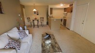 This single-floor residence features an open-concept layout that seamlessly connects the living room, dining area, and kitchen into one cohesive space. The living room is anchored by plush carpeting and accommodates comfortable seating alongside a granite-topped coffee table, creating a relaxed yet polished atmosphere. A warm neutral color palette runs throughout, with beige walls and light carpet contributing to the overall sense of openness.

The dining area is positioned adjacent to the living space and features a compact bistro-style table with tall bar chairs, illuminated by a hanging pendant light that adds a warm focal point overhead. A decorative accent wall in a deeper tan tone adds subtle visual contrast to the space.

The kitchen is defined by a peninsula counter that serves as both a workspace and a natural divider between the cooking and living areas. Medium-toned wood cabinetry lines the upper walls, and white appliances — including a refrigerator, stove, and microwave — provide a clean, functional look. The overall design balances practicality with a welcoming ambiance, making the space well-suited for everyday living.