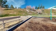 Located in Palmdale, California, this outdoor recreational space serves as a community amenity set against a natural hillside landscape. The grounds feature a vibrant, multi-component playground equipped with slides, climbing structures, and interactive play elements, all set within a wood chip safety surface and bordered by raised edging. Adjacent to the playground is a spacious paved court area with a basketball hoop and colorful line markings for a variety of games, accommodating both organized sports and casual play.

Picnic tables are positioned throughout the grounds, providing dedicated seating areas ideal for gatherings and outdoor dining. The open paved surfaces are marked with multiple court layouts in bright colors, maximizing the versatility of the space for different recreational activities. Tall light poles are distributed across the area, ensuring usability during evening hours.

A natural hillside rises along the perimeter, dotted with native shrubs, pine trees, and a palm tree, lending a scenic and open-air character to the environment. Benches are placed near the hillside edge, offering a quiet spot to rest with views of the surrounding landscape. The residential buildings nearby feature Spanish-style tile roofing and stucco exteriors. Ample parking is accessible on-site, and the overall layout reflects a well-organized, community-focused outdoor environment suited for residents of all ages.