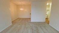 This single-floor space presents an open, understated layout characterized by neutral tones and simple, functional design. The interior features wall-to-wall carpeting in a warm beige tone that extends across the main living and dining areas, lending a cohesive and comfortable feel underfoot. A partial dividing wall separates the two zones while maintaining an open, connected atmosphere between them.

The dining area is anchored by a delicate chandelier that adds a classic, decorative touch to the otherwise minimalist space. Complementing this, a track lighting fixture runs along the ceiling of the adjacent living area, offering flexible, directed illumination throughout the room. The walls are finished in a soft, warm white that reflects light evenly and enhances the sense of openness.

Both areas benefit from clean baseboards and smooth wall surfaces, suggesting a well-maintained interior ready for personalization. A built-in feature, possibly a fireplace surround or recessed niche, is visible along one wall, adding a subtle architectural detail. Overall, the space offers a calm, move-in-ready environment with a straightforward layout well-suited for everyday living.