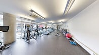 Located at 110 Winooski Falls Way in Winooski, Vermont, this fitness center is a well-equipped exercise space designed to support a well-rounded workout routine. The room features durable rubber flooring throughout, providing a safe and functional surface for a variety of exercises. Large floor-to-ceiling windows line one wall, flooding the space with natural light and offering views of the surrounding Winooski streetscape, creating an open and energizing atmosphere.

The cardio section is prominently featured, with a row of Matrix treadmills and elliptical trainers positioned along the windowed wall to take full advantage of the natural light and outdoor views. A stationary spin bike provides an additional cardio option. On the opposite side of the room, a multi-tiered dumbbell rack offers a comprehensive range of free weights, complemented by exercise balls, foam rollers, and yoga mats for stretching and core work. Full-length mirrors are mounted along one wall, enhancing the sense of space and allowing users to monitor their form. The room is illuminated by recessed and pendant fluorescent lighting, and a ceiling-mounted HVAC unit ensures climate control year-round. A frosted-glass entry door provides a clean, modern entryway into the facility.