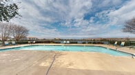 This single-story community amenity space is designed to offer both recreational and social functionality in an open, outdoor-oriented setting. The centerpiece is a generously sized swimming pool with clear blue water, surrounded by a broad concrete deck that provides ample space for lounging. The pool area is enclosed by a wrought-iron perimeter fence, beyond which expansive waterfront views stretch out beneath wide, open skies, creating a scenic backdrop that enhances the overall atmosphere.

Adjacent to the pool is a covered patio area supported by substantial textured columns with an arched architectural design. The structure features a ceiling fan overhead, providing comfort during warmer months. The covered space is furnished with modular outdoor seating, including a sectional sofa and coordinating accent chairs arranged around a low coffee table, creating a comfortable gathering area. The exterior walls of the structure are finished in a distinctive blue and cream color scheme, accented by wall-mounted lantern-style lighting fixtures and framed window panels.

Access to the pool area is controlled through a decorative iron gate set within an arched entryway, reinforcing the community's cohesive architectural style. The overall ambiance is relaxed and communal, with the waterfront views and open sky serving as defining features of the space.