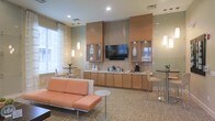 Located at 1982 Route 70 East in Cherry Hill, New Jersey, this single-floor commercial space offers a thoughtfully designed interior that balances functionality with a warm, inviting aesthetic. The lounge area serves as the centerpiece of the space, featuring a plush sectional sofa in a soft terracotta hue complemented by modern accent chairs and sleek side tables with chrome accents. The flooring is covered in a neutral, patterned carpet that adds texture while maintaining a professional atmosphere. A built-in cabinetry wall finished in warm wood tones houses a wall-mounted flat-screen television as the focal point, flanked by glass-front upper cabinets and a continuous lower cabinet run that doubles as a beverage and refreshment station. A decorative tile backsplash adds visual interest behind the cabinetry wall. Recessed lighting throughout the ceiling, paired with elegant pendant lights, ensures the space is well-lit and inviting. Natural light filters in through sheer-curtained windows, enhancing the bright and airy feel of the interior. High-top bistro tables with bar-height seating are positioned near the windows, providing additional gathering spots. The overall ambiance is polished, comfortable, and ideal for hospitality or professional use.