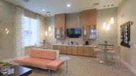 Located at 1982 Route 70 East in Cherry Hill, New Jersey, this single-floor commercial space offers a thoughtfully designed interior that balances functionality with a warm, inviting aesthetic. The lounge area serves as the centerpiece of the space, featuring a plush sectional sofa in a soft terracotta hue complemented by modern accent chairs and sleek side tables with chrome accents. The flooring is covered in a neutral, patterned carpet that adds texture while maintaining a professional atmosphere. A built-in cabinetry wall finished in warm wood tones houses a wall-mounted flat-screen television as the focal point, flanked by glass-front upper cabinets and a continuous lower cabinet run that doubles as a beverage and refreshment station. A decorative tile backsplash adds visual interest behind the cabinetry wall. Recessed lighting throughout the ceiling, paired with elegant pendant lights, ensures the space is well-lit and inviting. Natural light filters in through sheer-curtained windows, enhancing the bright and airy feel of the interior. High-top bistro tables with bar-height seating are positioned near the windows, providing additional gathering spots. The overall ambiance is polished, comfortable, and ideal for hospitality or professional use.