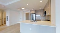 This single-floor space showcases a thoughtfully designed open-concept layout that seamlessly integrates the kitchen and living areas. The kitchen is a standout feature, anchored by a generous white quartz waterfall island that provides ample workspace and houses an undermount sink with a modern faucet. Upper cabinetry in a warm taupe finish with minimalist hardware lines the walls, complemented by a neutral-toned tile backsplash that adds subtle texture. Stainless steel appliances, including a French door refrigerator and built-in microwave, reinforce the kitchen's contemporary aesthetic. Recessed pot lighting throughout ensures bright, even illumination, while a flush-mount ceiling fixture adds a secondary light source in the adjacent area. Light-toned engineered hardwood flooring runs throughout, contributing to a clean and cohesive look. The entry area, visible from the kitchen, features a warm wood-toned door surround that introduces a natural element to the otherwise crisp, neutral palette. The overall ambiance is modern, bright, and functional, reflecting a refined yet understated design sensibility suited to contemporary living.