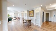 Located at 2500 Kelly Rd in Warrington, PA, this single-floor common area space offers a generously sized and thoughtfully designed environment suited for gathering and socializing. The interior is anchored by warm-toned hardwood flooring, which transitions between classic straight-lay and striking herringbone patterns depending on the zone, adding visual depth throughout.

The welcoming entrance features glass-paneled double doors with transom windows that flood the foyer with natural light. Wainscoting panels line the walls throughout, lending a refined, traditional character to every area. A reception-style lobby area includes built-in white shelving, ornate framed artwork, and a classic wood desk, establishing an inviting first impression.

The open kitchen is outfitted with warm wood cabinetry, white quartz countertops, glass-front upper cabinets, a double sink, and a stainless steel French door refrigerator. Adjacent to the kitchen, a buffet-style counter with matching cabinetry and a wall-mounted flat-screen TV provides a practical serving and entertainment hub.

The dining area accommodates a large white communal table with upholstered chairs beneath a statement globe chandelier and a coffered ceiling. Large windows offer views of the surrounding landscape. A comfortable lounge area with plush seating, curated artwork, and a chandelier-lit corridor leading to an elevator completes this polished, community-centered space.