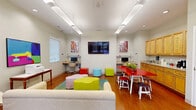 Located at 7798 Enfield Ave in Norfolk, Virginia, this bright and lively community room offers a thoughtfully designed, multi-functional space on a single floor. The interior is finished with warm hardwood flooring throughout and illuminated by a combination of recessed lighting and sleek linear pendant fixtures that keep the space feeling open and well-lit.

The lounge area anchors the room with a comfortable cream sofa, modern accent chairs, and a colorful area rug layered with playful modular seating pieces in bold hues of green, yellow, and red. A wall-mounted flat-screen TV serves as a central focal point, flanked by vibrant artwork that adds energy and personality to the neutral walls.

A bright red dining table with white chairs and stools provides a casual gathering spot near the kitchenette, which features warm wood cabinetry, a sink, and counter space — ideal for light food and beverage preparation. Two built-in computer workstations with granite-topped desks are tucked along the walls, offering functional workspace within the communal setting.

Large glass doors along one wall flood the space with natural light and open to an outdoor patio area, enhancing the airy atmosphere. A glimpse through an interior glass partition reveals an adjacent fitness area, adding to the suite of amenities available within this dynamic community space.