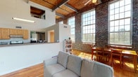 This two-story loft showcases a striking industrial character rooted in its original architectural bones. The main living level is anchored by soaring ceilings with warm wood-beam detailing and exposed steel structural elements, creating a dramatic sense of volume and openness. Original exposed brick walls dominate the space, particularly in the living and dining areas, where floor-to-ceiling factory-style windows flood the interior with abundant natural light and offer views of the surrounding landscape.

The open-concept living area features hardwood floors throughout, with the dining space nestled into a sunlit brick corner framed by tall multi-pane windows. The kitchen is positioned on an elevated pass-through counter arrangement with light wood cabinetry, granite-style countertops, and standard appliances, open to both the living and dining areas.

A wood-tread staircase with white risers connects the main floor to the upper level, where the bedrooms are located. Bedrooms feature carpeted floors and exposed beam ceilings, with windows overlooking exterior brick details. One bedroom on the lower level also features exposed structural ceiling elements. The bathrooms are cleanly appointed, with one featuring a pedestal sink and shower-tub combination. The overall ambiance is warm, open, and rich in industrial-loft character.