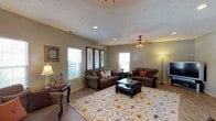 This single-story home presents a welcoming and well-appointed living space designed with comfort and functionality in mind. The living room serves as the focal point of the home, showcasing a vaulted ceiling that adds a sense of openness and volume to the space. A textured ceiling detail adds subtle architectural character, while recessed lighting and a ceiling fan provide both practical illumination and air circulation.

The room is finished with warm wood-style flooring that runs throughout, grounding the space with a natural, inviting aesthetic. Neutral sage-green walls create a calming backdrop that complements the rich, dark wood tones of the furniture. Multiple windows allow generous natural light to flow into the room, enhancing the bright and airy atmosphere.

The living area is generously proportioned, comfortably accommodating two substantial upholstered sofas arranged around a dark wood coffee table atop a large decorative area rug. Framed botanical artwork adorns the walls, adding a refined touch. A floor lamp and table lamps contribute warm ambient lighting, creating a cozy, layered effect. The overall ambiance is warm, casual, and well-balanced, ideal for both everyday living and entertaining.