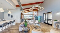 This single-floor community room in Westlake, Louisiana, delivers a well-appointed shared gathering space defined by striking architectural character and versatile amenities. Soaring ceilings with exposed rustic wood beams and an industrial-style ductwork detail lend the main lounge area a modern yet warm aesthetic. A dramatic ring-shaped pendant light fixture serves as a central focal point overhead, while wide-plank wood-look flooring runs throughout, tying the space together cohesively.

The expansive lounge area is furnished with plush sofas, accent chairs, and glass-topped coffee tables arranged to encourage both social interaction and relaxed lounging. Large floor-to-ceiling windows flood the interior with natural light and provide open views of the surrounding exterior, enhancing the airy atmosphere. A wall-mounted television is positioned over a wood-paneled accent wall, adding a media-ready component to the seating area.

The kitchen features a substantial white quartz island with bar seating, soft blue cabinetry, a matching subway tile backsplash, and pendant lighting overhead. A wall-mounted television, sink, and stainless steel appliances make it fully functional for gatherings and events.

A dedicated game area houses a full-sized billiards table bathed in natural light from an expansive glazed window wall. A pet-supply vending machine near the entry adds a unique, community-friendly amenity to this thoughtfully designed space.