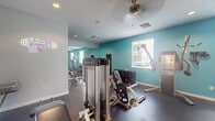 This fitness center is a thoughtfully designed, single-level space that offers a comprehensive workout environment. The interior is characterized by a two-tone color scheme, combining crisp white walls with vibrant teal accent walls that create an energetic and motivating atmosphere. Dark rubberized flooring spans the entire area, providing both durability and safety underfoot, while a textured ceiling with recessed lighting and ceiling fans ensures consistent air circulation and illumination throughout.

The space is divided into distinct functional zones. The strength training area features a robust selection of Freemotion cable machines, a leg press, and a fully stocked dumbbell rack with a medicine ball station. Floor-to-ceiling mirrors line one wall, visually expanding the space and allowing users to monitor their form. A neon motivational sign adds a modern, stylish touch to this section.

The cardio zone is equipped with treadmills, elliptical machines, and a stationary bike, each positioned near wall-mounted televisions for entertainment during workouts. Multiple windows fitted with blinds allow natural light to filter in while maintaining privacy. A dedicated exit door provides convenient access. Overall, the space delivers a clean, functional, and energizing fitness experience.