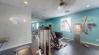 This fitness center is a thoughtfully designed, single-level space that offers a comprehensive workout environment. The interior is characterized by a two-tone color scheme, combining crisp white walls with vibrant teal accent walls that create an energetic and motivating atmosphere. Dark rubberized flooring spans the entire area, providing both durability and safety underfoot, while a textured ceiling with recessed lighting and ceiling fans ensures consistent air circulation and illumination throughout.

The space is divided into distinct functional zones. The strength training area features a robust selection of Freemotion cable machines, a leg press, and a fully stocked dumbbell rack with a medicine ball station. Floor-to-ceiling mirrors line one wall, visually expanding the space and allowing users to monitor their form. A neon motivational sign adds a modern, stylish touch to this section.

The cardio zone is equipped with treadmills, elliptical machines, and a stationary bike, each positioned near wall-mounted televisions for entertainment during workouts. Multiple windows fitted with blinds allow natural light to filter in while maintaining privacy. A dedicated exit door provides convenient access. Overall, the space delivers a clean, functional, and energizing fitness experience.