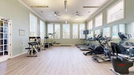 Located at 2800 W Baker Rd in Baytown, Texas, this fitness center offers a spacious, single-floor exercise facility designed to support a well-rounded workout experience. The room is finished with light hardwood-style flooring that lends a clean, modern aesthetic throughout the open layout.

Tall, white-trimmed windows line multiple walls, flooding the space with abundant natural light and offering views of the surrounding outdoor greenery, including palm trees and neighboring structures. The walls are painted in a soft sage green, creating a calm and refreshing atmosphere conducive to exercise.

The ceiling is notably elevated, featuring elegant crown molding and dual ceiling fans that promote airflow and comfort during workouts. Large wall-mounted mirrors are strategically placed to maximize the sense of openness and allow users to monitor their form.

The facility is comprehensively equipped with multiple treadmills, elliptical machines, stationary bikes, and a rowing machine for cardiovascular training. Strength equipment includes a multi-function cable machine, a leg press/extension machine, adjustable benches, and a dumbbell rack. A wall-mounted flat-screen TV adds an entertainment element for cardio sessions. The space is accessed through elegant French doors with glass panels, reinforcing the refined architectural character of the facility.