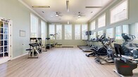Located at 2800 W Baker Rd in Baytown, Texas, this fitness center offers a spacious, single-floor exercise facility designed to support a well-rounded workout experience. The room is finished with light hardwood-style flooring that lends a clean, modern aesthetic throughout the open layout.

Tall, white-trimmed windows line multiple walls, flooding the space with abundant natural light and offering views of the surrounding outdoor greenery, including palm trees and neighboring structures. The walls are painted in a soft sage green, creating a calm and refreshing atmosphere conducive to exercise.

The ceiling is notably elevated, featuring elegant crown molding and dual ceiling fans that promote airflow and comfort during workouts. Large wall-mounted mirrors are strategically placed to maximize the sense of openness and allow users to monitor their form.

The facility is comprehensively equipped with multiple treadmills, elliptical machines, stationary bikes, and a rowing machine for cardiovascular training. Strength equipment includes a multi-function cable machine, a leg press/extension machine, adjustable benches, and a dumbbell rack. A wall-mounted flat-screen TV adds an entertainment element for cardio sessions. The space is accessed through elegant French doors with glass panels, reinforcing the refined architectural character of the facility.
