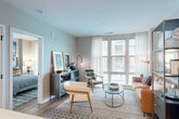This thoughtfully designed one-bedroom, one-bathroom residence offers a contemporary open-concept layout that maximizes every square foot. The living room serves as the heart of the space, featuring large floor-to-ceiling windows dressed with sheer curtains that flood the room with natural light and provide views of the surrounding cityscape. Light wood flooring flows seamlessly throughout, complementing the neutral, soft gray tones on the walls.

The kitchen is both functional and elegant, boasting a two-tone cabinet design with upper white cabinetry and lower gray units, paired with sleek quartz countertops, a tile backsplash, and a full suite of stainless steel appliances including a refrigerator, electric range, and microwave. A kitchen island with an undermount sink adds prep space and connects the kitchen visually to the living area.

The bedroom is a bright and restful retreat, with oversized windows that continue the theme of natural light. It accommodates a dedicated workspace alongside the sleeping area, and includes a closet for ample storage.

The bathroom features large-format gray tile, a deep soaking tub with an overhead shower, matte black fixtures, and a clean, minimalist aesthetic. A natural wood entry door with modern hardware completes this well-appointed residence.