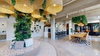 Located at 2585 Spruce Ave in West Palm Beach, Florida, this thoughtfully designed single-floor commercial space offers a generous and open layout that seamlessly blends nature-inspired aesthetics with modern functionality. The entrance welcomes visitors with ornate patterned tile flooring, a striking terrazzo-topped reception desk, and a moss-lettered logo wall accented by a neon "Come As You Are" sign — establishing a warm, inclusive atmosphere from the moment of arrival. Oversized rattan pendant lights and trailing greenery further reinforce the space's vibrant, biophilic design language. The open main area flows into a collaborative work zone featuring a long communal wooden table lined with amber upholstered chairs, set beneath a dramatic overhead installation of cascading foliage and Edison bulb pendants. Floor-to-ceiling black-framed windows flood the interior with abundant natural light and frame views of mature tropical palms outside. A lounge nook with a sleek channeled sofa and sculptural marble-topped tables provides an inviting area for informal gatherings. A glass-enclosed private meeting room offers a quieter setting for focused work. A bar-style counter with patterned tile detailing and black barstools, paired with a gallery wall of gold-framed mirrors and potted greenery, completes this eclectic and energizing space.
