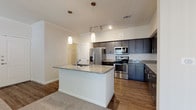This single-floor, one-bedroom, one-bathroom residence offers a thoughtfully designed layout with modern finishes throughout. The heart of the home is a well-equipped kitchen featuring rich, dark-toned cabinetry with sleek hardware, granite countertops, and a full suite of stainless steel appliances including a refrigerator with ice maker, microwave, and range. A central kitchen island with a granite surface and integrated sink serves as both a functional workspace and a natural gathering point, illuminated by elegant pendant lighting above. Wood-look flooring runs throughout the kitchen and main areas, creating a warm, cohesive aesthetic. Track lighting complements the pendant fixtures, ensuring the kitchen is well-lit from multiple angles. The living area transitions seamlessly from the kitchen, featuring neutral carpet flooring and a ceiling fan for added comfort. The bedroom is a calm, carpeted retreat with a ceiling fan, natural light from a window with blinds, and ample space. The bathroom is clean and bright, with white tile surrounding a full soaking tub and shower combo, wood-look flooring, and modern brushed nickel fixtures. A walk-in closet is accessible from the main hallway, adding convenient storage to this efficiently designed home.