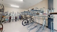 Located at 1505 Water Tower Cir in Greenville, South Carolina, this dedicated indoor bicycle storage room offers a well-organized, secure space for cyclists. The room spans a generous single-floor footprint, providing ample capacity for numerous bicycles across a thoughtfully designed layout.

The space is equipped with multiple rows of galvanized steel two-tier bicycle racks, maximizing vertical storage and allowing a high volume of bikes to be stored efficiently. Both the upper and lower tiers feature individual wheel cradles and support arms designed to securely hold a variety of bicycle styles, from road bikes to mountain bikes.

Overhead, flush-mounted fluorescent lighting ensures the entire room is brightly and evenly illuminated. The flooring is a smooth, durable light gray surface that is easy to maintain. The walls are finished in a neutral off-white tone, contributing to a clean and functional atmosphere.

Access to the storage room is controlled via a secured door with an electronic keypad entry system, ensuring only authorized users can enter. A set of arched glass double doors at one end of the room allows for easy bicycle roll-in and roll-out, with views of the surrounding greenery beyond. Decorative cycling-themed artwork is displayed on the walls, adding a welcoming touch to this practical amenity.