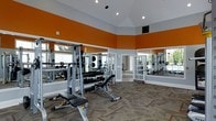 This fitness center is designed to provide a motivating and functional workout environment. The space is defined by its striking vaulted ceiling, which creates an airy, open atmosphere and adds a sense of grandeur to the room. Bold orange accent walls contrast sharply with the light gray upper walls and white trim, lending the space an energetic and modern aesthetic.

The flooring features a patterned carpet that adds texture and comfort underfoot, while large floor-to-ceiling mirrors line multiple walls, visually expanding the space and allowing users to monitor their form during exercise. Recessed lighting is integrated into the ceiling, ensuring the room is well-lit throughout.

The facility is outfitted with a comprehensive selection of strength training equipment, including a multi-functional cable and weight machine, an adjustable workout bench, and a full rack of free weights and dumbbells. The equipment is thoughtfully arranged to allow for comfortable movement and accessibility throughout the space.

Natural light filters in through windows and glass-paneled French doors, which also provide views of and access to the surrounding exterior. The overall ambiance is clean, bright, and well-maintained, making this fitness center a practical and inviting space for a full range of workout routines.