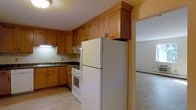 This single-floor residence offers a functional and comfortable layout designed for everyday living. The kitchen serves as a central feature of the home, outfitted with warm-toned wood cabinetry that extends to the ceiling, maximizing storage space. Dark stone countertops provide a practical workspace, complemented by a full suite of white appliances including a gas range with oven and a full-size top-freezer refrigerator. An under-cabinet light fixture illuminates the workspace, while a sink positioned along the side wall adds to the kitchen's efficiency. The cabinetry's classic raised-panel style and crown molding detailing lend a traditional character to the space. The kitchen flows openly into an adjacent room featuring light-colored carpeting, which provides a softer, more relaxed atmosphere. A baseboard heating unit along the wall of the adjoining room ensures year-round comfort. The warm yellow-toned walls throughout create a welcoming ambiance, tying the spaces together cohesively. The overall layout is straightforward and practical, offering a comfortable living environment with well-considered details throughout.