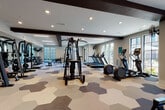 This expansive fitness center is designed to accommodate a wide range of workout styles within a single, thoughtfully organized space. The interior is anchored by a striking hexagonal-patterned floor in neutral tones of cream, gray, and blue, providing both visual interest and functional zoning throughout the facility. Dark exposed beams span the white ceiling, adding architectural contrast and a modern industrial character to the open layout.

Large black-framed windows line multiple walls, flooding the space with natural light and offering views of the surrounding exterior. Recessed lighting and linear LED fixtures supplement illumination throughout. A ceiling fan enhances airflow in key areas of the room.

The equipment layout is comprehensive, featuring treadmills, ellipticals, stationary bikes, cable machines, a stair climber, free weights, kettlebells, battle ropes, plyometric boxes, and a rowing ergometer. A mirrored free-weight zone with a weight bench allows for strength training. An open stretch and functional training area provides ample floor space for bodyweight or accessory exercises.

A dedicated amenity nook finished with glossy dark subway tile houses a water fountain, rolled towel service, yoga mats, and storage cubbies, adding a polished, spa-like convenience to the facility. A reclaimed wood accent wall near the entrance further enhances the upscale aesthetic of this well-appointed fitness space.