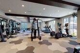 This expansive fitness center is designed to accommodate a wide range of workout styles within a single, thoughtfully organized space. The interior is anchored by a striking hexagonal-patterned floor in neutral tones of cream, gray, and blue, providing both visual interest and functional zoning throughout the facility. Dark exposed beams span the white ceiling, adding architectural contrast and a modern industrial character to the open layout.

Large black-framed windows line multiple walls, flooding the space with natural light and offering views of the surrounding exterior. Recessed lighting and linear LED fixtures supplement illumination throughout. A ceiling fan enhances airflow in key areas of the room.

The equipment layout is comprehensive, featuring treadmills, ellipticals, stationary bikes, cable machines, a stair climber, free weights, kettlebells, battle ropes, plyometric boxes, and a rowing ergometer. A mirrored free-weight zone with a weight bench allows for strength training. An open stretch and functional training area provides ample floor space for bodyweight or accessory exercises.

A dedicated amenity nook finished with glossy dark subway tile houses a water fountain, rolled towel service, yoga mats, and storage cubbies, adding a polished, spa-like convenience to the facility. A reclaimed wood accent wall near the entrance further enhances the upscale aesthetic of this well-appointed fitness space.