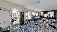 This single-level fitness center offers a comprehensive workout environment designed with both functionality and aesthetics in mind. The main exercise floor is generously sized, accommodating a wide array of cardio equipment including treadmills, ellipticals, and stair climbers arranged in rows facing large windows that frame views of the surrounding community, including palm trees and a pool area, flooding the space with natural light.

A dedicated free weights section features a full rack of dumbbells displayed against a wall accented with warm wood-toned paneling and a large mirror, which visually expands the space and supports strength training. A Hoist cable machine and additional resistance equipment are positioned nearby for versatile training options.

A separate spin studio houses several cycling bikes and is equipped with a wall-mounted flat-screen television, a mirrored wall with matching wood-framed accents, and recessed lighting, creating an energizing atmosphere for cycling sessions.

Throughout the facility, the flooring consists of patterned carpet tiles in a blue-grey herringbone style, adding a modern, polished look. Recessed lighting brightens the interior uniformly, while black-framed glass doors connect the fitness center to an adjacent outdoor patio area, promoting a seamless indoor-outdoor flow.
