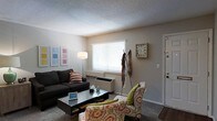 This single-floor space offers a comfortable and functional layout designed for everyday living. The living room serves as the heart of the home, featuring neutral beige walls and a textured ceiling that add a sense of warmth and character. A large window dressed with white horizontal blinds allows natural light to fill the room, creating a bright and inviting atmosphere. The space is thoughtfully arranged with a dark sectional sofa accented by striped throw pillows, a sleek glass-top coffee table, and a patterned accent chair in earthy tones with a green throw pillow, adding a pop of color and personality. A freestanding floor lamp provides additional ambient lighting, enhancing the room's cozy feel. A wall-mounted clock and a set of framed wall art contribute to the room's curated, homey aesthetic. A coat rack near the entryway offers practical storage upon arrival. The flooring transitions between carpet in the living area and what appears to be hard flooring near the entry, adding visual definition to the space. The front door is positioned conveniently off the living area, making for an efficient and welcoming flow throughout this charming single-floor home.