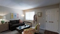 This single-floor space offers a comfortable and functional layout designed for everyday living. The living room serves as the heart of the home, featuring neutral beige walls and a textured ceiling that add a sense of warmth and character. A large window dressed with white horizontal blinds allows natural light to fill the room, creating a bright and inviting atmosphere. The space is thoughtfully arranged with a dark sectional sofa accented by striped throw pillows, a sleek glass-top coffee table, and a patterned accent chair in earthy tones with a green throw pillow, adding a pop of color and personality. A freestanding floor lamp provides additional ambient lighting, enhancing the room's cozy feel. A wall-mounted clock and a set of framed wall art contribute to the room's curated, homey aesthetic. A coat rack near the entryway offers practical storage upon arrival. The flooring transitions between carpet in the living area and what appears to be hard flooring near the entry, adding visual definition to the space. The front door is positioned conveniently off the living area, making for an efficient and welcoming flow throughout this charming single-floor home.