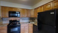 Located at 1700 Street Road in Warrington, Pennsylvania, this single-floor residence offers a functional and thoughtfully designed living space. The kitchen stands out as a central feature, showcasing rich, honey-toned raised-panel wood cabinetry that extends to the upper walls, providing generous storage throughout. A decorative mosaic stone tile backsplash adds texture and visual interest, complementing the granite-style countertops below. The kitchen is equipped with a full suite of black appliances, including a gas range with overhead microwave, a dishwasher, and a full-size refrigerator, creating a cohesive and practical cooking environment. A corner sink with a single-basin design maximizes counter space while maintaining an efficient workflow. The layout makes smart use of every corner, offering an organized and approachable kitchen suited for everyday use. The overall design balances warm, natural tones with classic finishes, giving the space an inviting and comfortable character consistent throughout the single-level floor plan.
