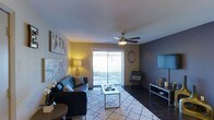 This single-story residence offers three bedrooms and two bathrooms within a thoughtfully arranged floor plan. The living room serves as the central gathering space, featuring a ceiling fan with integrated lighting and angled walls that add subtle architectural character. A sliding glass door floods the room with natural light and provides direct access to an outdoor patio, creating a seamless indoor-outdoor connection. The contrasting wall tones—warm yellow and deep charcoal gray—lend the space a modern, intentional aesthetic. Durable dark flooring runs throughout, complemented by a neutral area rug that defines the seating area. Vertical blinds on both the sliding door and an adjacent window offer flexible light control and privacy. The open arrangement of the living area allows for versatile furniture placement, accommodating both relaxed lounging and casual entertaining. With three bedrooms, the residence provides ample flexibility for use as sleeping quarters, a home office, or a guest room. The two full bathrooms add convenience for residents and guests alike. Overall, the layout is practical and well-suited for shared or family living.