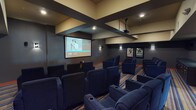This private screening room is designed to replicate the full cinematic experience in a refined, intimate setting. The space features rows of plush, navy blue theater-style armchairs arranged in a gently tiered layout, each equipped with built-in cupholders for added comfort during screenings. A ceiling-mounted projector beams onto a large, centrally positioned projection screen at the front of the room, providing clear, expansive viewing from every seat.

The architectural design draws attention upward with a striking coffered ceiling featuring bold, dark geometric beams set against a warm tan background, adding depth and a dramatic visual character to the room. The walls are finished in deep charcoal, accented by warm wall sconces that provide subtle ambient lighting. Classic framed movie posters line the side walls, reinforcing the cinematic theme throughout.

A striped carpet in tones of blue, tan, and brown runs the length of the room, adding color and visual warmth underfoot. Toward the rear of the space, a popcorn machine and a small concession counter complete the theater experience. A vibrant red door at the back provides a bold accent and serves as the main entry point. The overall ambiance is immersive, stylish, and purpose-built for an exceptional private movie-watching experience.