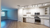 This well-appointed single-floor residence offers a clean, contemporary aesthetic throughout. The kitchen serves as a functional centerpiece, featuring crisp white shaker-style cabinetry with brushed nickel hardware, dark countertops, a tile backsplash, and a full suite of stainless steel appliances including a gas range, microwave, dishwasher, and refrigerator. Track lighting runs along the ceiling, providing excellent task illumination across the workspace. Upper cabinets include glass-front display panels, adding a refined detail to the kitchen design. Light-toned hardwood flooring extends seamlessly through the kitchen and into the open living areas, creating a bright and cohesive feel. The living space is generously proportioned, with neutral-toned walls, clean white baseboards, and a door that opens to a windowed exterior entry. A hallway connects the main living area to the private quarters, where a walk-in closet provides substantial storage with multiple chrome hanging rods and shelving on two walls over plush carpeted flooring. The layout flows intuitively, and the consistent use of light tones, modern finishes, and thoughtful design details gives the space a fresh, move-in-ready character throughout.