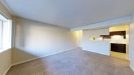 This single-floor residence offers a clean and functional layout designed for comfortable everyday living. The main living area is spacious and open, finished with wall-to-wall neutral-toned carpeting that lends a warm, understated feel to the room. Smooth, light-colored walls and a textured ceiling contribute to a bright and airy atmosphere, while white baseboards provide a crisp finishing detail throughout.

A defining architectural feature is the pass-through opening connecting the living area to the kitchen, which allows for natural sightlines between the two spaces and promotes an open, connected feel. The kitchen is partially visible through this opening, suggesting a compact yet practical cooking area. A doorway at the far end of the space provides access to additional rooms or the unit entrance, contributing to a logical and easy-to-navigate floor plan.

Overhead lighting in the transition area between the living room and kitchen provides warm illumination, complementing the natural light in the space. The overall ambiance is neutral and move-in ready, offering a blank canvas that can be easily personalized. The single-floor layout ensures convenience and accessibility throughout, making it a practical and welcoming living environment.