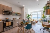 This single-floor residence offers a thoughtfully designed open-concept layout that seamlessly connects the kitchen, dining, and living areas. The kitchen is outfitted with stainless steel appliances—including a range, microwave, dishwasher, and refrigerator—complemented by light wood cabinetry, a white tile backsplash, clean quartz countertops, and open metal shelving. Light wood-look flooring runs throughout, enhancing the airy, cohesive feel of the space.

The dining area sits adjacent to the kitchen and accommodates a round glass table, while the living area beyond is bathed in natural light from large floor-to-ceiling windows that frame stunning, unobstructed views of the beach and ocean. This sun-filled living space provides a serene and inviting atmosphere year-round.

The bedroom continues the beachfront theme, featuring a generous window with direct ocean and beach views, creating a tranquil retreat. Recessed lighting throughout the residence adds warmth and functionality.

The bathroom is clean and modern, featuring a floating wood-tone vanity with an illuminated mirror, a medicine cabinet, and neutral tile flooring, offering both practicality and style. Overall, the residence presents a bright, coastal-inspired ambiance with contemporary finishes throughout.