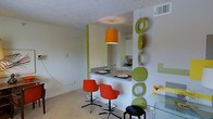 This single-floor space showcases a lively, retro-modern aesthetic defined by bold color choices and playful design elements. The open layout seamlessly connects the kitchen and dining areas, creating a cohesive and social environment. A striking orange pendant light serves as a focal point, casting warm light over the bar-style counter that separates the kitchen from the living and dining zones. The counter accommodates seating with a pair of vibrant red-orange modern bucket chairs on sleek metallic bases, complemented by a black stool, adding a dynamic mix of seating options.

A decorative wall installation featuring alternating lime-green rings and discs adds a whimsical, artistic element to the space, drawing the eye vertically and adding depth to the room. Lime-green accent columns and wall details echo this color scheme throughout. The kitchen, visible beyond the counter, features white cabinetry and a functional layout. Light-toned carpeting in the main area provides a neutral contrast to the bold accents. A glass-top table is partially visible, hinting at a dedicated dining area. The overall ambiance is energetic, creative, and distinctly retro-inspired.
