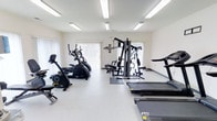 This fitness center offers a practical and well-organized workout environment designed to accommodate a range of exercise needs. The space is single-level and finished with clean white walls and a light-toned floor, creating a bright and open atmosphere that is both inviting and functional. Overhead fluorescent lighting ensures consistent, even illumination throughout the room, complemented by natural light filtering in through large windows dressed with vertical blinds along the far wall. The cardio section features a stationary exercise bike, an elliptical trainer, and a treadmill from the 6000 Series line, providing users with multiple low-impact and high-intensity options. A multi-station strength training machine occupies the center of the space, offering a versatile platform for resistance and weight-based workouts. Additional equipment is visible toward the rear of the room, further expanding the training possibilities available. The layout is thoughtfully arranged to allow comfortable movement between stations, and rubber floor mats are positioned beneath key equipment for safety and stability. Overall, the space presents a clean, accessible, and fully functional fitness facility suited for a broad range of users and workout routines.