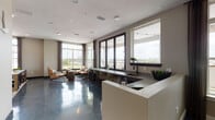 This contemporary single-floor amenity space is thoughtfully designed to blend functionality with modern aesthetics. The open-concept layout is anchored by striking polished concrete floors that reflect natural light streaming in through the expansive floor-to-ceiling and large-pane windows, which frame sweeping panoramic views of the surrounding skyline and landscape.

The main gathering area features a generous lounge zone furnished with a plush dark leather sectional sofa, accent chairs, and a low-profile coffee table set atop a layered area rug. Adjacent to this seating area is a dedicated dining nook with a round concrete-top table and modern wire chairs positioned beside a large window with access to an outdoor balcony, where string lights add a warm ambiance.

A built-in kitchen and bar counter runs along the window wall, complete with an undermount sink, dark countertops, and bar-height seating — ideal for casual gatherings or collaborative use. The kitchen area also includes a dedicated plant shelf and access to the outdoor terrace through glass-panel doors.

A distinct lounge alcove is defined by a reclaimed wood accent wall, a textured charcoal feature panel, and a wall-mounted television, creating an intimate media retreat. Curated artwork and sculptural wall décor are displayed throughout, contributing to the space's refined yet inviting character. Recessed ceiling lighting and integrated speakers complete the polished, modern environment.