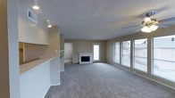 This single-floor residence offers a well-organized and functional layout designed for comfortable everyday living. The main living area is generously sized, featuring soft wall-to-wall carpeting that adds warmth underfoot and a neutral color palette throughout that creates a calm, cohesive atmosphere. A standout feature of the living room is the classic white-brick fireplace, which serves as a natural focal point and adds a cozy, inviting character to the space. Large windows along one wall allow natural light to fill the room, providing views of the surrounding exterior. A glass-paneled exterior door offers additional access and further enhances the sense of openness. The textured popcorn ceiling adds a traditional finish overhead, while track lighting near the entryway provides supplemental illumination. A half-wall partition separates the living area from an adjoining space, suggesting a semi-open floor plan that balances connectivity with defined zones. A hallway visible from the main room leads to additional areas of the home, indicating a thoughtful layout that prioritizes flow and accessibility. Overall, this space combines practical design with classic residential details, making it a solid and welcoming environment.
