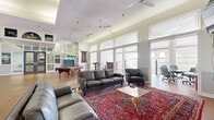 Located at 500 Tripoli Street in Pittsburgh, PA, this generously sized community floor offers an impressive blend of functionality and classic architectural character. The open layout is anchored by soaring ceilings — highlighted by a coffered skylight detail in the central gathering area — and warmed throughout by rich hardwood flooring. Rows of tall, white-trimmed windows flood the space with abundant natural light, while multiple ceiling fans maintain a comfortable atmosphere.

The main lounge area is furnished with plush leather sofas and armchairs, arranged atop ornate Persian-style area rugs, creating an inviting setting for socializing. A regulation pool table and a ping-pong table provide recreational options, complementing a mounted flat-screen TV for entertainment. A built-in bar with seating offers an additional social hub near the exterior access points.

A dedicated meeting room, visible through a wide doorway, features a round conference table and office-style seating. Floor-to-ceiling glass-panel partitions with steel-framed double doors define a corridor leading to adjacent building wings, maintaining visual openness while delineating zones. An ornate wrought-iron staircase with arched windows adds a refined, historic architectural touch. The overall ambiance is bright, welcoming, and thoughtfully designed for community engagement.