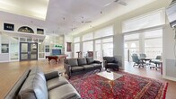 Located at 500 Tripoli Street in Pittsburgh, PA, this generously sized community floor offers an impressive blend of functionality and classic architectural character. The open layout is anchored by soaring ceilings — highlighted by a coffered skylight detail in the central gathering area — and warmed throughout by rich hardwood flooring. Rows of tall, white-trimmed windows flood the space with abundant natural light, while multiple ceiling fans maintain a comfortable atmosphere.

The main lounge area is furnished with plush leather sofas and armchairs, arranged atop ornate Persian-style area rugs, creating an inviting setting for socializing. A regulation pool table and a ping-pong table provide recreational options, complementing a mounted flat-screen TV for entertainment. A built-in bar with seating offers an additional social hub near the exterior access points.

A dedicated meeting room, visible through a wide doorway, features a round conference table and office-style seating. Floor-to-ceiling glass-panel partitions with steel-framed double doors define a corridor leading to adjacent building wings, maintaining visual openness while delineating zones. An ornate wrought-iron staircase with arched windows adds a refined, historic architectural touch. The overall ambiance is bright, welcoming, and thoughtfully designed for community engagement.