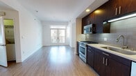 This contemporary single-floor unit showcases a clean, modern design with wood-tone flooring throughout the living areas and bedrooms. The open-concept layout connects the kitchen directly to the living space, creating a seamless, airy flow. The kitchen is well-appointed with rich dark-wood cabinetry extending floor to ceiling, light stone countertops, a gray tile backsplash, and a full suite of stainless steel appliances including a gas range, built-in microwave, dishwasher, and refrigerator. Recessed lighting highlights the kitchen's sleek finishes. The living area benefits from generous natural light and direct access to a private balcony through glass-paned French doors, offering views of surrounding greenery and the neighborhood beyond. The bedrooms feature hardwood-style flooring, large windows with tree-lined outdoor views, and built-in closets for ample storage. Two bathrooms serve the unit, each finished with light tile flooring and dark-wood vanities with white countertops. The primary bathroom includes a glass-enclosed walk-in shower, while the secondary bathroom offers a clean, functional layout with grab bars. A utility room with an electrical panel completes the unit's practical infrastructure. The overall aesthetic is bright and contemporary.