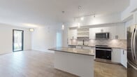 Located at 3991 NW 82nd Dr in Cooper City, Florida, this single-floor residence showcases a clean, modern aesthetic with an open-concept design that connects the living, dining, and kitchen areas seamlessly. Wood-look tile flooring flows consistently throughout the space, enhancing the bright, airy atmosphere created by abundant natural light from large windows and a glass-paneled entry door.

The kitchen is a standout feature, equipped with crisp white shaker cabinetry accented by matte black hardware, a striking chevron-patterned tile backsplash, and a full suite of stainless steel appliances including a French door refrigerator, range, microwave, and dishwasher. A large center island with a neutral stone-finish countertop provides additional prep space and casual seating. Pendant lighting and a track lighting system illuminate the area stylishly.

The generously sized living area offers plenty of natural light and includes a ceiling fan, while the bedrooms feature wide windows with views of the surrounding greenery.

The bathrooms are finished to a high standard, with wood-look tile flooring, white cabinetry with matte black hardware, and frameless glass walk-in showers clad in large-format neutral tiles. The overall design palette — whites, warm wood tones, and soft grays — creates a cohesive, contemporary atmosphere throughout.