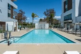 Located at 2277 Harbor Blvd in Costa Mesa, California, this outdoor amenity deck is a well-appointed resort-style pool area set within a modern residential community. The centerpiece is a generously sized rectangular swimming pool with crystal-clear blue water, surrounded by a spacious sun deck finished in light-toned concrete pavers that reflect the abundant Southern California sunshine. Adjacent to the main pool is a spa equipped with an ADA-compliant mechanical pool lift, ensuring accessible entry for all residents. The deck is thoughtfully furnished with a variety of chaise lounges and seating arrangements, both in open sun and beneath pergola-style shade structures. These white-framed pergolas with slatted roofs are positioned along the pool perimeter, providing comfortable shaded retreats. The surrounding architecture features a contemporary multi-story building clad in crisp white stucco and warm wood-toned panels, with glass-railed balconies overlooking the pool area. Lush tropical landscaping, including palm trees and ornamental plantings, frames the space and reinforces the Southern California aesthetic. The pool area benefits from open sky exposure, allowing for ample natural light throughout the day, making it an inviting outdoor destination for relaxation and recreation.